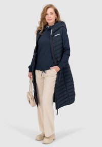 Gilet lungo imbottito di colore blu navy con cappuccio, indossato sopra una felpa blu navy e pantaloni beige. Gli accessori includono una piccola borsa beige e scarpe chunky.