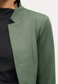 Άτομο που φοράει υφασμάτινο σακάκι ελιάς πάνω από ένα μαύρο τοπ, το οποίο είναι κομμένο ώστε να φαίνεται ο ώμος και η περιοχή του άνω στήθους.