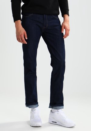 Persona con vaqueros azul oscuro remangados, calcetines blancos, zapatillas Nike blancas y una camiseta negra de manga larga, de pie sobre un fondo claro.