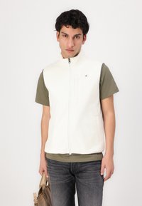 Weißer Fleece-Weste mit hohem Kragen, Frontreißverschluss und Seitentaschen, kombiniert mit einem grünen kurzärmeligen Hemd und dunkler Jeans.