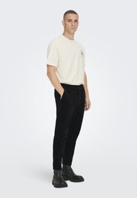 Beige Baumwoll-T-Shirt mit Rundhalsausschnitt und Logo auf der linken Brust, kombiniert mit schwarzen Cordhosen und schwarzen Stiefeletten.