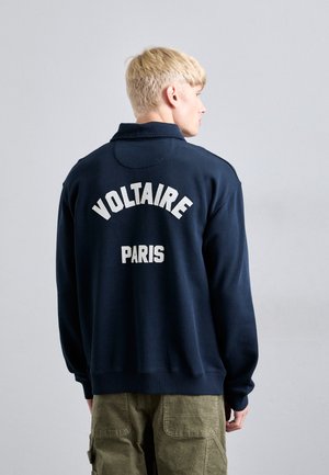 Blond person facing away wearing a navy blue sweatshirt with "VOLTAIRE" and "PARIS" in white text on the back, and olive green pants.