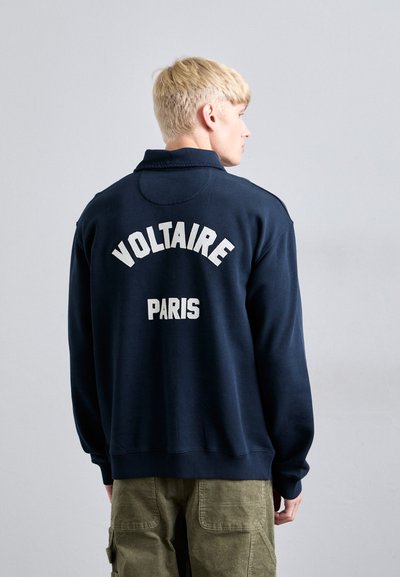 En blond person, der vender væk, iført en marineblå sweatshirt med "VOLTAIRE" og "PARIS" i hvid tekst på ryggen, samt olivengrønne bukser.