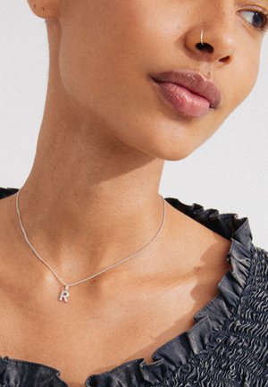 Collana d'argento con un piccolo pendant a forma di "R", indossata su pelle nuda, abbinata a un top di tessuto scuro, strutturato e arricciato.