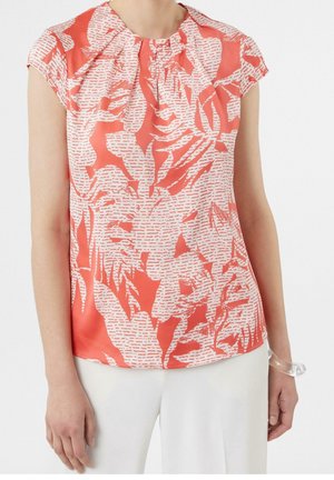 Femme portant un chemisier sans manches corail rose avec des motifs abstraits de feuilles blanches et un pantalon blanc sur un fond uni.