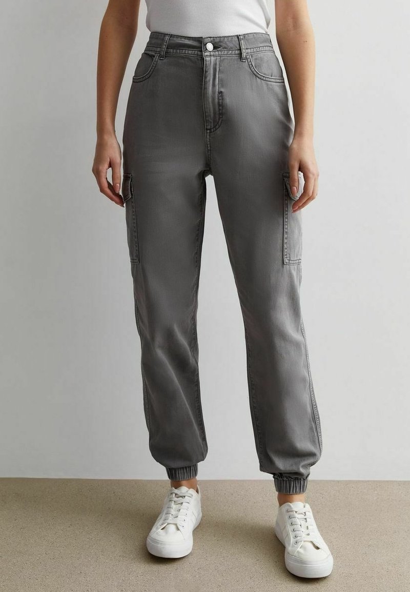 Personne portant un pantalon cargo gris taille haute avec des bracelets élastiques et des baskets blanches, debout devant un fond clair uni.