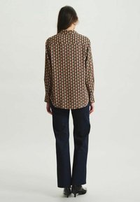 Blouse à manches longues avec un motif géométrique rouge, noir et beige, associée à un jean bleu foncé et des chaussures à talons noirs.