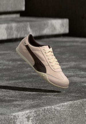 Zapatilla deportiva beige y marrón con cordones suspendida sobre escalones de hormigón texturizado con sombra debajo.