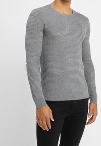 Gray knitted long-sleeve sweater with a textured pattern, round neckline, and ribbed cuffs. Paired with dark pants for a contrasting look.
