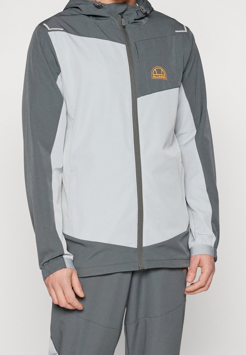Veste gris clair et gris foncé en tissu respirant, avec fermeture éclair frontale, capuche et une poche poitrine avec un logo.