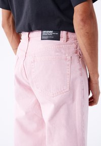 Vaqueros de mezclilla rosa claro con un corte relajado, con bolsillos traseros y una etiqueta de marca negra en la cinturilla. Textura suave, sin patrones visibles.