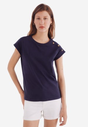 Navy cotton t-shirt with short sleeves and a rounded neckline, featuring three gold buttons on the left shoulder, paired with white shorts.