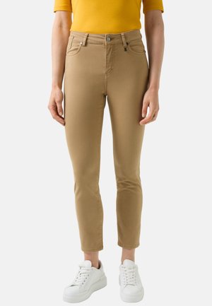 Frau trägt beige enge Hose, gelbes kurzärmeliges Oberteil und weiße Turnschuhe, steht vor einem schlichten hellen Hintergrund.