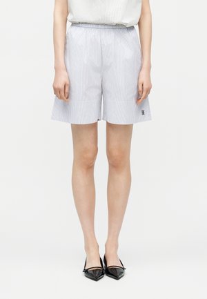 Osoba nosząca jasnoniebieskie spodenki w pionowe paski z elastycznym pasem oraz czarne, szpiczaste baleriny, stojąca na białym tle.