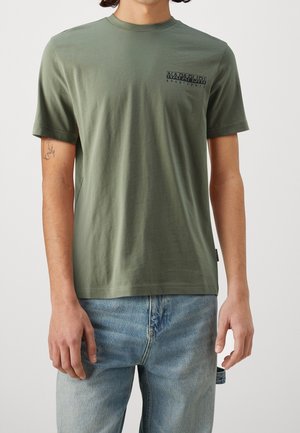 Uomo che indossa una t-shirt Napapijri verde oliva a maniche corte con piccolo logo sul petto e jeans azzurri chiari contro uno sfondo neutro.