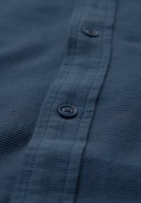 Granatowy, teksturowany materiał z poziomymi paskami i detalem guzika, z wyraźnym guzikiem z wygrawerowanym logo.