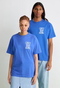 Zilā kokvilnas T-krekli ar apaļu kakla izgriezumu. Priekšā rakstīts "OBEY World Wide Records" baltā krāsā. Modelis pa kreisi valkā džinsa svārkus.