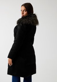 Cappotto imbottito nero con colletto in pelliccia, caratterizzato da un design trapuntato e vita stretta, che arriva fino a metà coscia. Texture liscia e vestibilità sagomata.