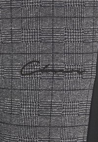 Texturerad tyg med ett mönster av marinblå och silverrutigt, med en upphöjd svart broderad logotypdetalj på ena sidan.