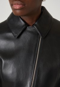Černá kožená bunda s ostrým límcem a stříbrným zipem. Textura je hladká, s viditelným šitím a strukturovaným designem.