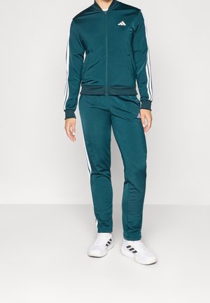 Personne portant un survêtement Adidas vert foncé avec des bandes blanches sur les manches et les jambes, associé à des baskets blanches avec des bandes noires.