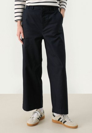 Personne portant un pantalon noir à jambes larges, des baskets blanches avec des bandes noires, des chaussettes blanches, et une chemise blanche à manches longues avec des rayures horizontales noires.