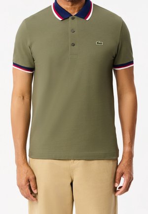 Homme portant un polo vert olive avec un col et des bordures de manches bleu marine, rouge et blanc, associé à un pantalon beige, debout devant un fond blanc.