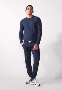 Navy blue sweat set made of soft material; features a crew neck sweatshirt and drawstring pants with pockets. Silver-accented sneakers complete the look.
