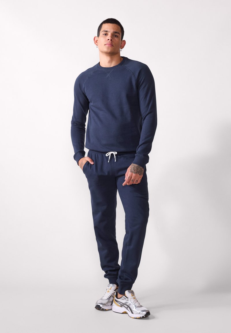 Navy blue sweat set made of soft material; features a crew neck sweatshirt and drawstring pants with pockets. Silver-accented sneakers complete the look.