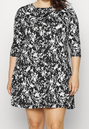 Robe noire et blanche à motif abstrait, longueur au genou, avec manches trois quarts, portée par une personne aux cheveux bruns.