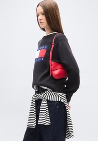 Jeune femme portant un sweat Tommy Jeans noir, un jean foncé, une chemise rayée nouée à la taille, et un sac à épaule rouge, debout devant un fond uni.