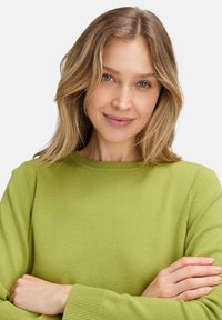 Zöld kötött pulóver kerek nyakkivágással és bordás mandzsettával, puha textúrával és testhezálló fazonnal. A modellen keresztbefont karokkal áll.