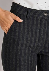 Hand met lichtroze nagels rustend in de zak van donkergestreepte stoffen broek met een knoop bij de taille.