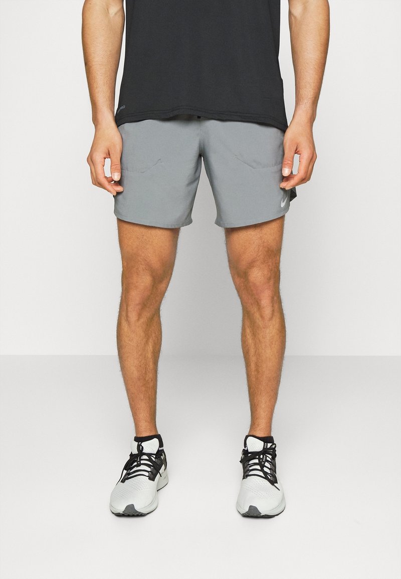 Homme portant un short de sport gris, un haut noir et des chaussures de course blanches, debout devant un fond uni.