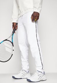 Vita sportbyxor med svarta sidostråk, i kombination med en vit texturerad jacka och vita sneakers med blå detaljer, medan man håller en tennisracket.