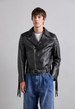 Jachetă de motocicletă din piele neagră, cu fermoare, talie cu curea și accente franjurate pe mâneci. Pur și simplu cu pantaloni din denim ușor albastri.
