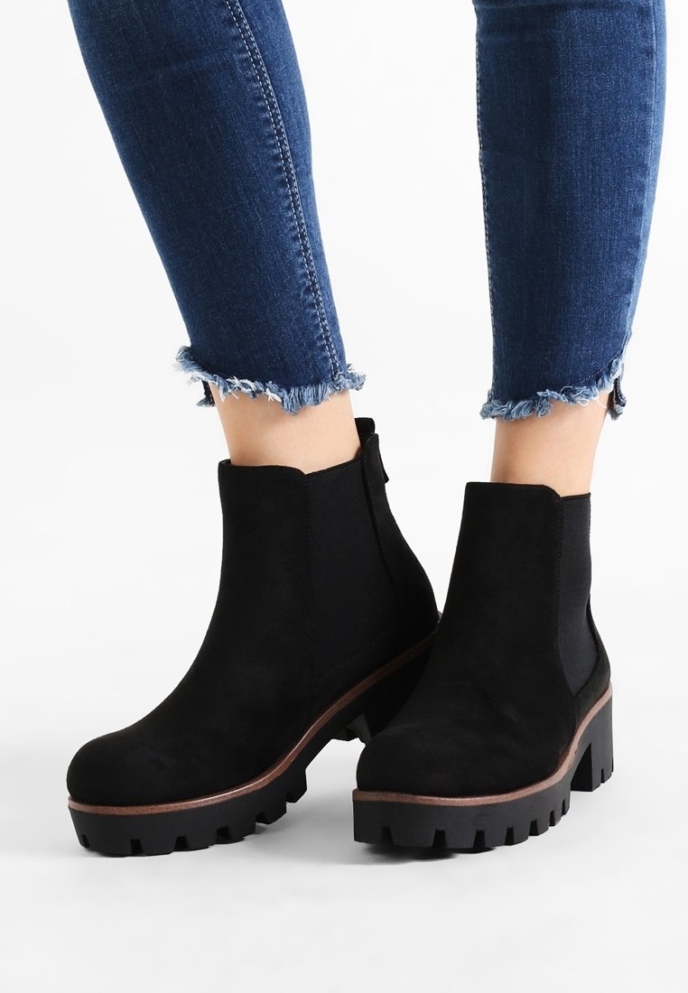 Bottes cheville en daim noir avec semelles crantées épaisses portées avec un jean skinny bleu à ourlets effilochés sur fond blanc.