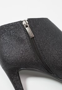Scarpa nera con tacco alto caratterizzata da una texture brillantinata, dettagli con zip sul lato e un design elegante con punta affilata.