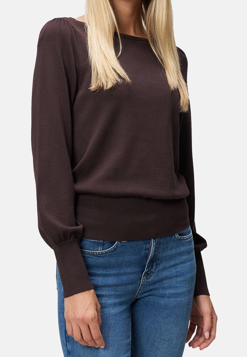 Femme portant un pull à manches longues marron foncé avec des épaules bouffantes et des manches retroussées, associé à un jean bleu.