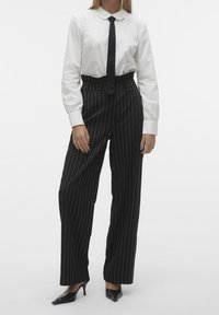 Chemise blanche à col, cravate noire, pantalon noir taille haute avec des rayures verticales blanches, tissu lisse, et escarpins noirs pointus.