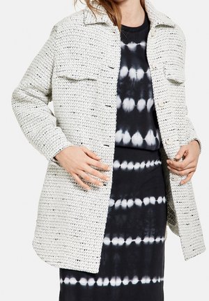 Nainen, jolla on valkoinen teksturoitu napillinen takki mustavalkoisen, solmuvärjätyn mekon päällä, kädet säätelemässä takin etuosaa.