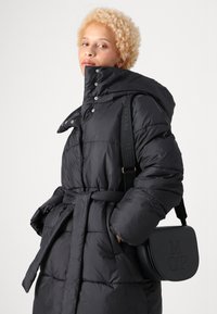 Giubbotto imbottito nero con colletto alto, bottoni a scatto e vita con cintura. Presenta una texture liscia. La modella tiene una borsa a tracolla nera.