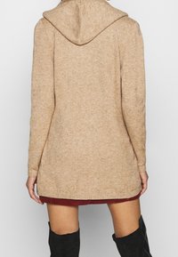 Béžový měkký pletený cardigan s kapucí, dlouhými rukávy a mírně prodlouženým střihem. Má žebrované manžety a lem.