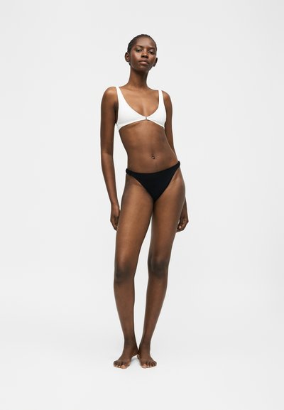 Γυναίκα που στέκεται ξυπόλητη φορώντας λευκό τοπ μπικίνι και μαύρο κάτω μέρος μπικίνι μπροστά σε απλό ανοιχτό γκρι φόντο.