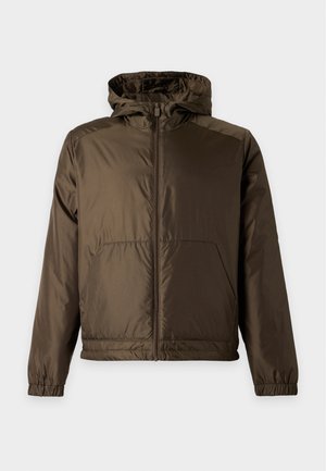 Homme debout portant une veste zippée vert olive, chemise blanche, pantalon noir et chaussures noires sur un fond clair uni.