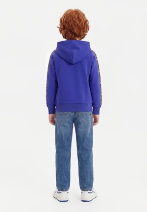 Barn med krøllet rødt hår iført en blå hættetrøje, blå jeans og hvide sneakers, der står med ryggen til mod en ensfarvet hvid baggrund.