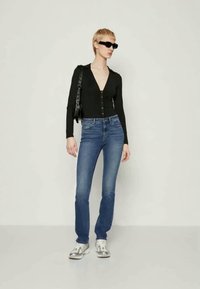 Μαύρη μακρυμάνικη μπλούζα με κουμπιά, μπλε τζιν και ασημένια αθλητικά παπούτσια; το μοντέλο ποζάρει με μια μαύρη τσάντα και μαύρα γυαλιά ηλίου.