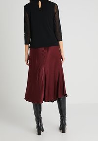 Haut noir avec des manches transparentes trois-quarts et un détail en trou de serrure au dos, associé à une jupe en satin bordeaux et des bottes noires jusqu'aux genoux.