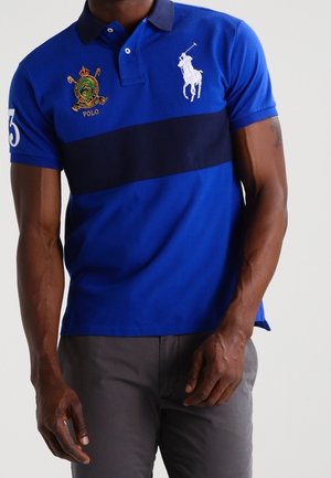 Mann trägt ein blaues Poloshirt mit dunklem horizontalem Streifen, besticktem weißem Polo-Spieler-Logo und Wappenemblem auf der Brust, kombiniert mit grauen Hosen.