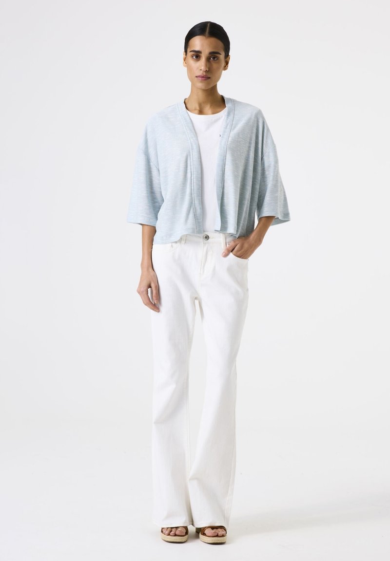 Lichtblauwe cropped jas met een open voorkant, brede mouwen en een zachte textuur, gecombineerd met witte flared broek en bruine plateausandalen.